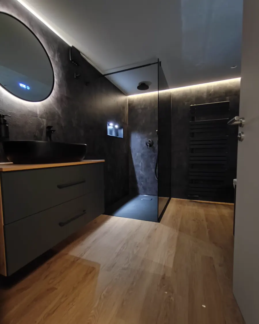 Badezimmer mit rundem hinterleuchtetem Spiegel, schwarzem Aufsatzbecken auf Holzplatte, bodengleicher Dusche hinter Glas und schwarzem Handtuchheizkörper.