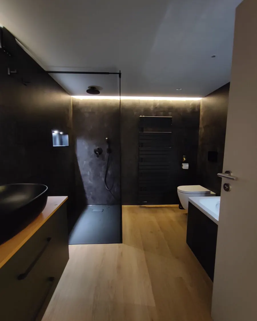 Das Bad von der Tür aus: Dusche, WC, Heizkörper und Holzboden unter einer umlaufenden Lichtvoute.