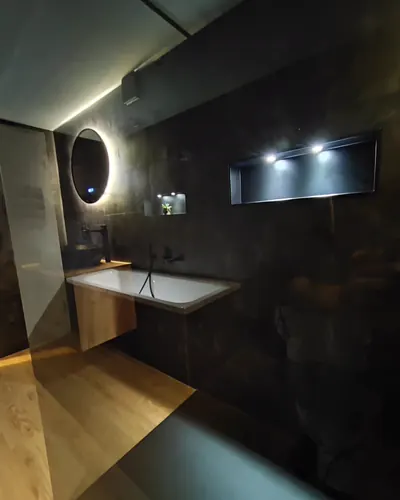 Badewanne mit Holzverkleidung unter einer langen beleuchteten Wandnische, daneben ein runder hinterleuchteter Spiegel.