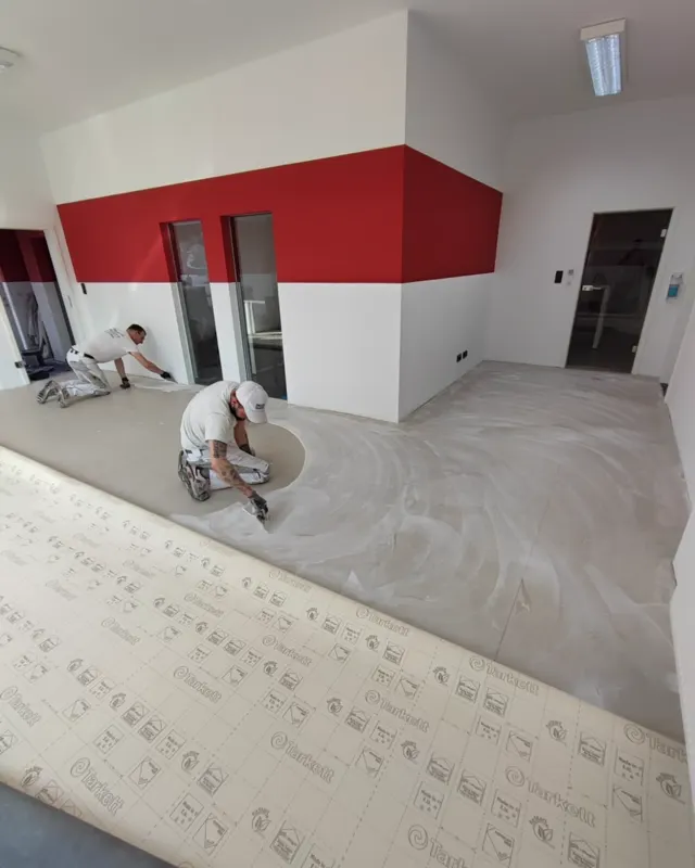 Zwei Mitarbeiter beim Spachteln des Bodens in einer Gewerbefläche mit rot-weiß gegliederter Wand