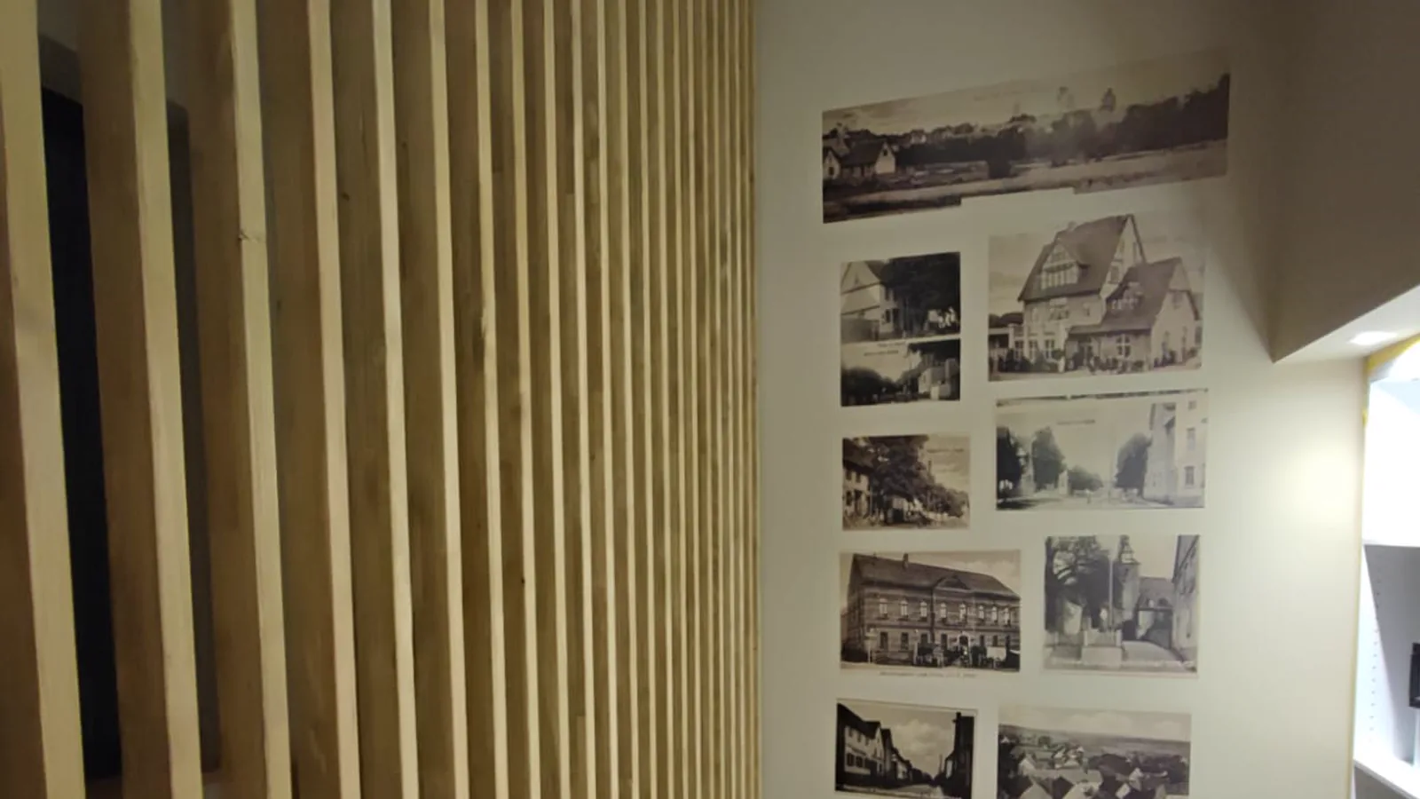 Raum mit einer raumhohen Wand aus senkrechten Eichenlamellen; rechts daneben eine Wand, auf die historische Ortsansichten in Sepia gedruckt sind.
