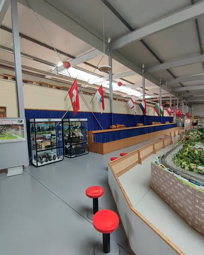 Blick in die Halle mit leerer heller Wand über der Vorhangreihe, davor rote Sitzhocker und eine Modellbahnanlage.