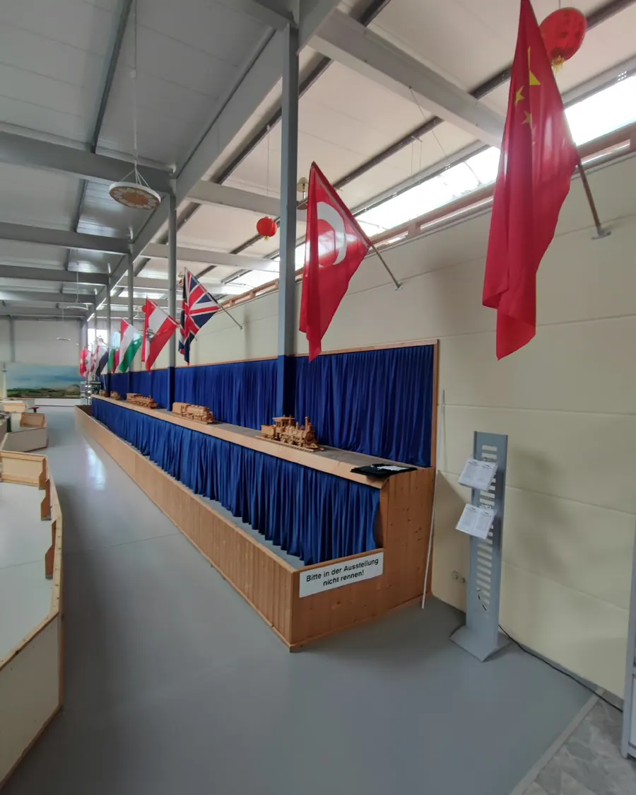 Ausstellungshalle mit cremefarbener Wand über einer blauen Vorhangreihe, darüber hängen Flaggen von der Hallendecke.