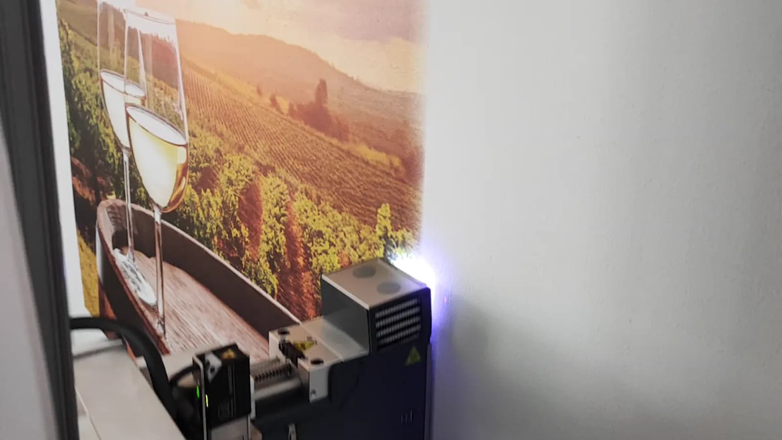 Der Druckkopf des Wanddruckers steht an einer Wand und trägt ein Weinberg-Motiv mit zwei Weißweingläsern auf. Am Kopf leuchtet die violette Trocknerlampe.