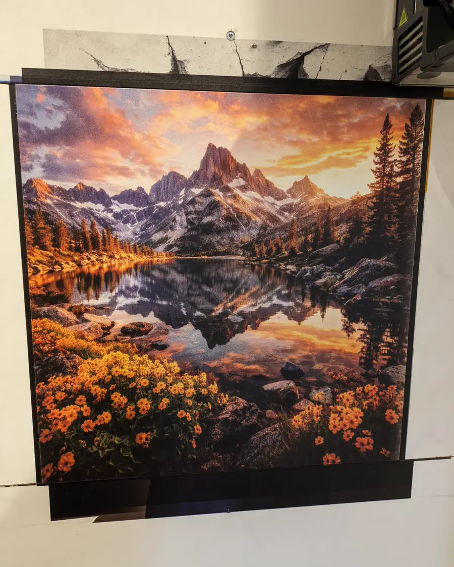 Bedruckte Platte mit einem Bergsee im Abendlicht, umgeben von Nadelwald und orangefarbenen Blumen. Oben rechts der Druckkopf.