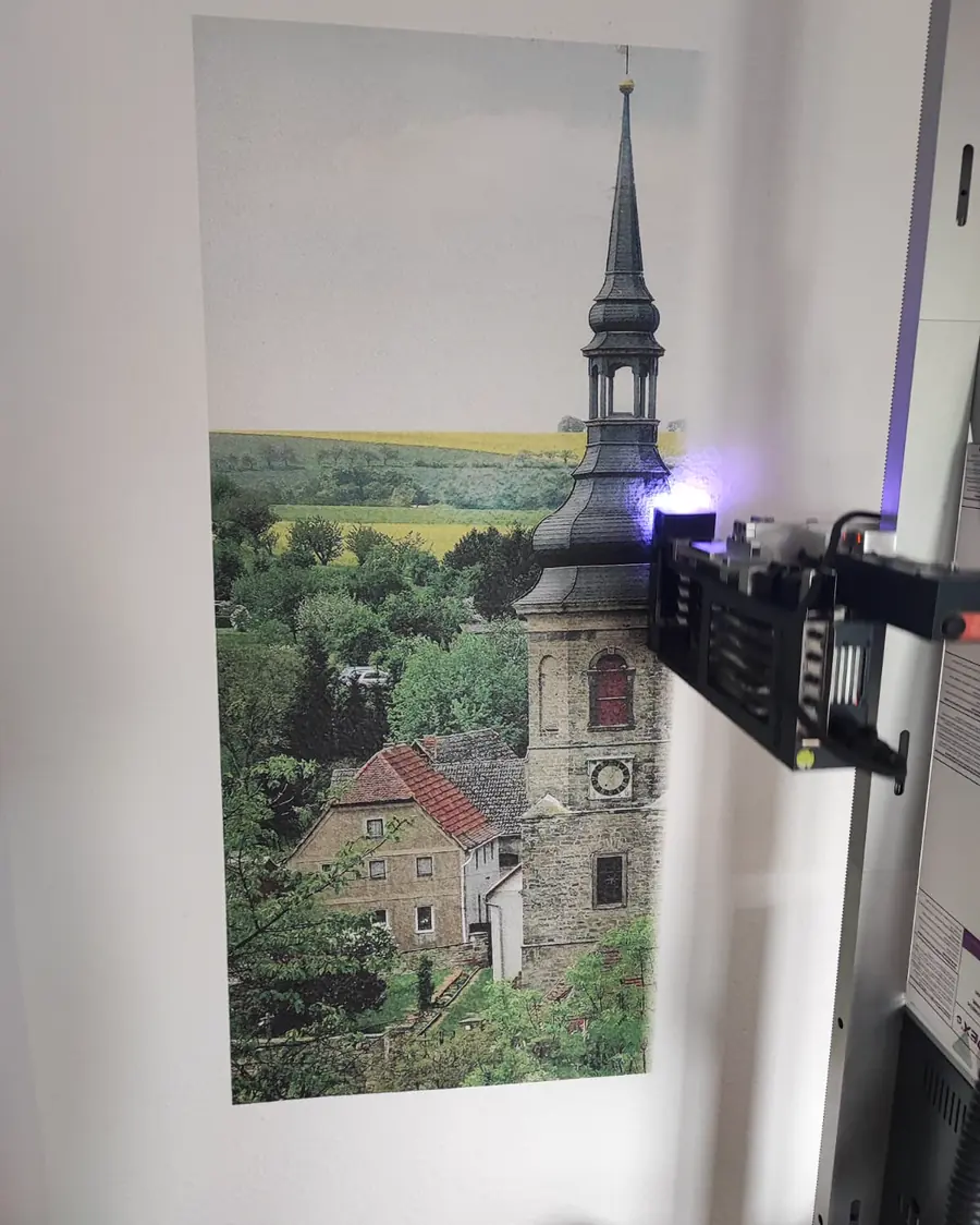 Der Druckkopf trägt ein Motiv auf eine Wand auf: eine Dorfkirche mit Zwiebelturm vor Rapsfeldern. Am Kopf leuchtet die violette Trocknerlampe.