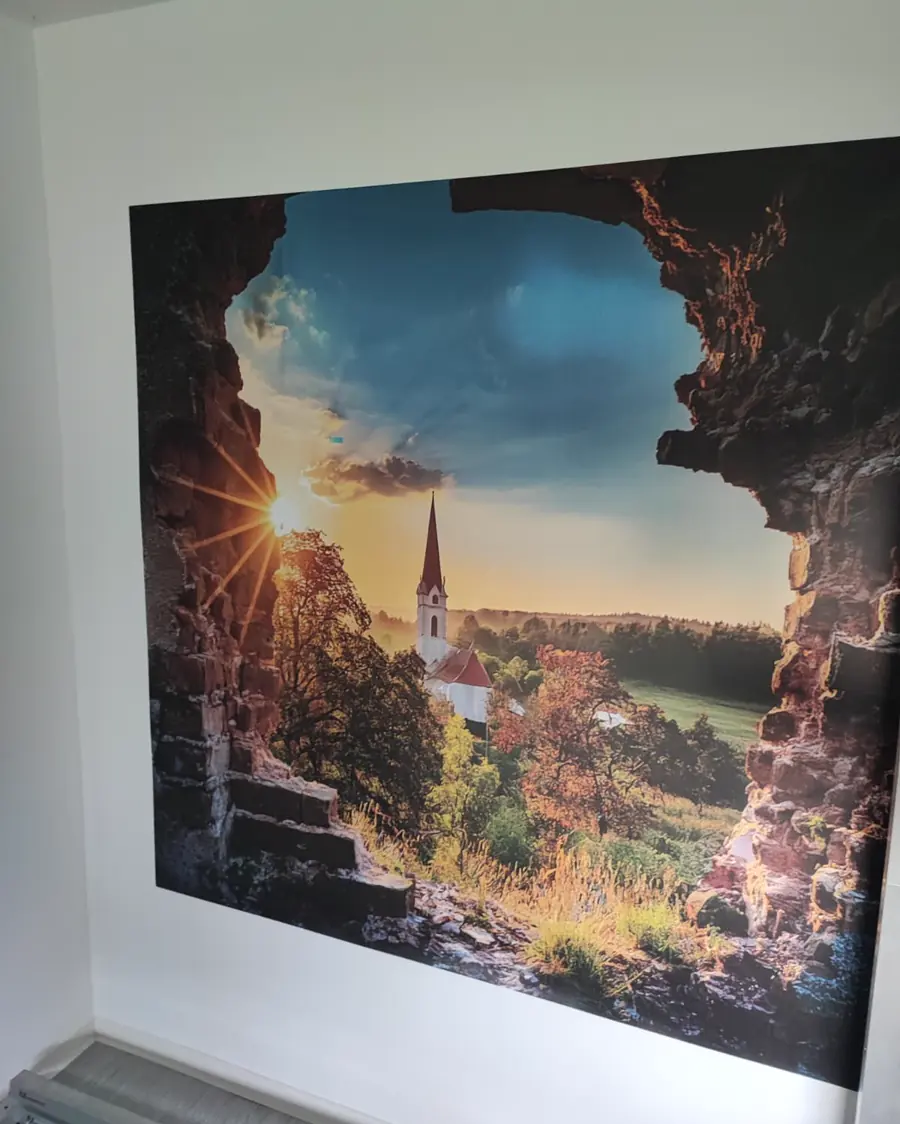 Wandbild, das einen Durchbruch in einer Ruinenmauer zeigt: dahinter eine Dorfkirche im Gegenlicht der untergehenden Sonne.